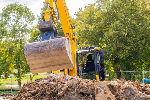 An excavator on a construction site lifts soil near trees and a cloudy sky, building a PV open air
