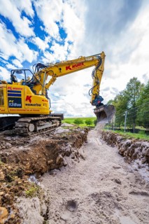 A yellow excavator carries out trenching work on a construction site under a cloudy sky, Bau PV