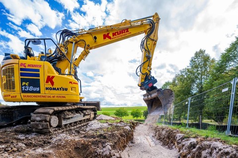 A yellow excavator digging on a construction site under a cloudy sky, Bau PV Freifaechenanlage,