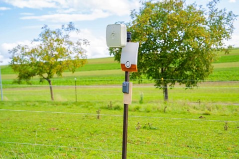 Camera on a post, surrounded by green fields and trees, against a cloudy sky, Bau PV