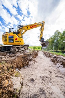 A yellow excavator in a deep ditch surrounded by nature under a cloudy sky, construction PV