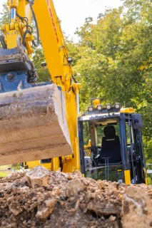 Close-up of an excavator with a raised grab arm at a construction site in front of trees, PV