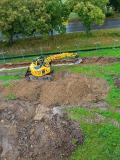 Excavator on a construction site with excavated soil surrounded by greenery and trees, Bau PV