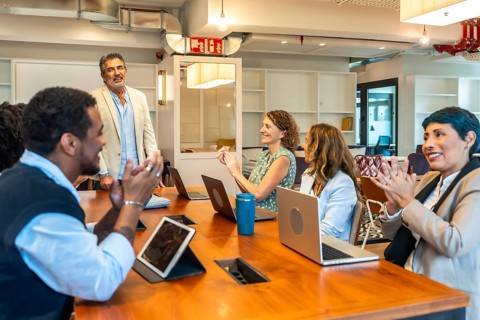 Diverse business colleagues sitting around a large wooden table, smiling and clapping for a speaker