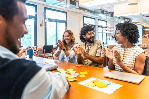 Diverse business colleagues sitting at a table in a modern office, happily clapping and smiling