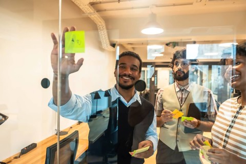 Happy diverse business team collaborating on glass wall during brainstorming session, sharing ideas