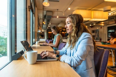 Woman with a headset and microphone actively working on a laptop at a long table by a window,