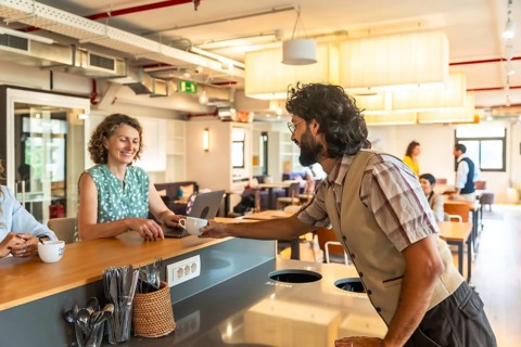 Young man passing a cup of coffee to a smiling woman working on her laptop at a counter in a