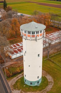 Close-up of a water tower surrounded by trees with autumn leaves, next to buildings with red roofs,