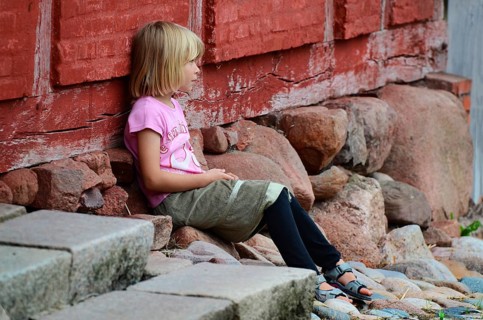 Little blonde girl sitting next to a red half-timbered house and looking to the side in Den gamle