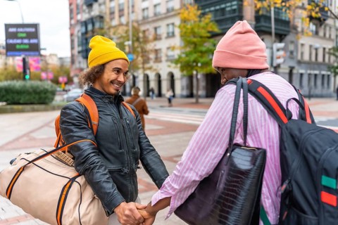 Multiethnic gay couple holding hands, beanies and backpacks, smiling as they walk through a vibrant