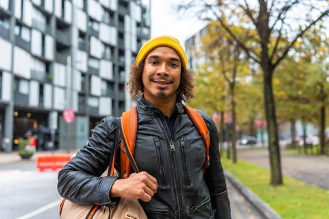 Young man with curly hair in a yellow beanie and leather jacket, backpack on his shoulders, smiling