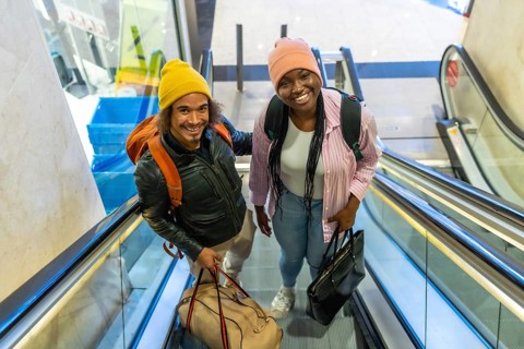 Happy young couple moving up an escalator at an airport or train station, carrying their travel