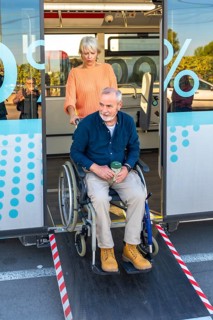 Senior woman is confidently assisting a mature man in a wheelchair to exit a public bus using the