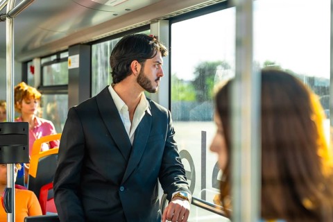 Young businessman in a dark suit stands on a city bus, gazing out the window as warm sunlight