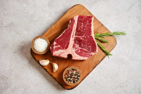 A large, marbled raw t-bone steak on a wooden cutting board surrounded by garlic cloves, salt,