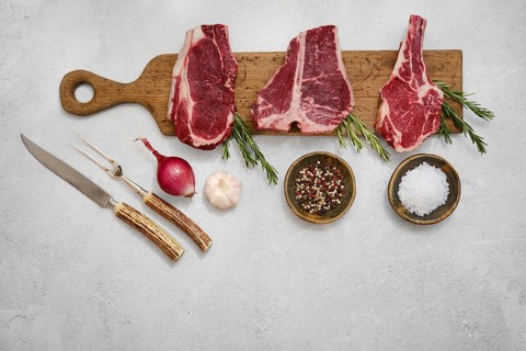 Three thick cuts of beef steaks on a wooden chopping board with fresh rosemary, salt, pepper, onion