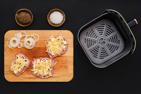 Raw pork chops are topped with grated cheese and sliced onions on a wooden cutting board. Nearby,