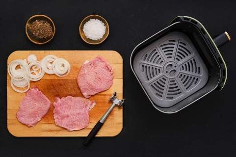 Raw pork chops on a wooden cutting board next to mallet. Sliced onions are ready beside them, with
