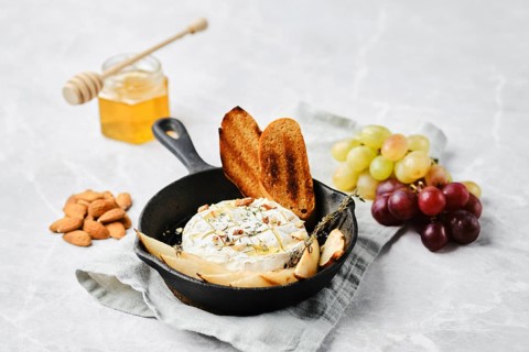 A cast iron skillet holds warm baked cheese, garnished with herbs. Nearby are toasted bread slices,
