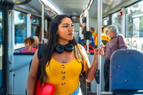 Young woman passenger holding a handrail inside a public bus, wearing headphones and a backpack,