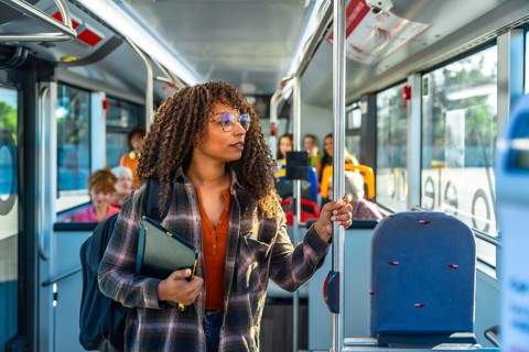 Young woman with curly hair and glasses stands inside a city bus holding a pole, wearing a backpack