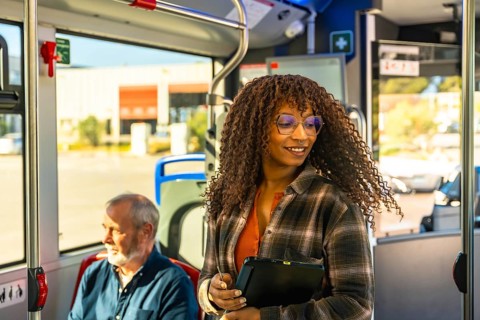 Woman with glasses smiles while standing on a city bus, holding a digital tablet among commuters