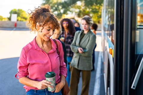 Smiling woman holding a reusable coffee cup waits in line at a busy city bus stop with diverse
