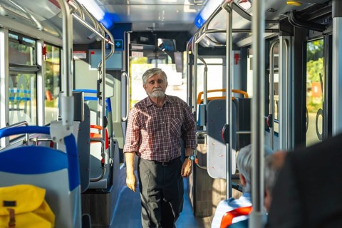 Senior man with a beard and plaid shirt standing in the aisle of a public bus, commuting during the
