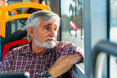 Senior man with gray hair and beard traveling on a public bus, wearing a plaid shirt and looking