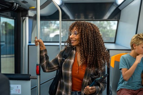 Woman with glasses and curly hair holding a digital tablet while commuting on a public bus, smiling