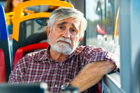 Senior man with white beard and plaid shirt sits by a bus window, gazing pensively at the camera