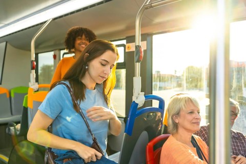 Diverse commuters on a city bus in sunlight, young woman looking down while other passengers sit