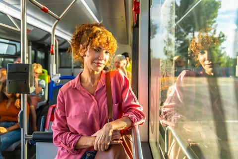Young redheaded woman smiling and looking at camera while standing on a city bus, holding a bag