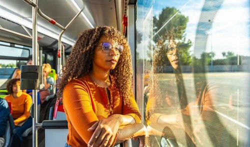 Young adult woman with curly hair and glasses looking out a public transport bus window, reflecting