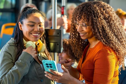 Two smiling multiethnic women bonding and sharing content on a smartphone while commuting together