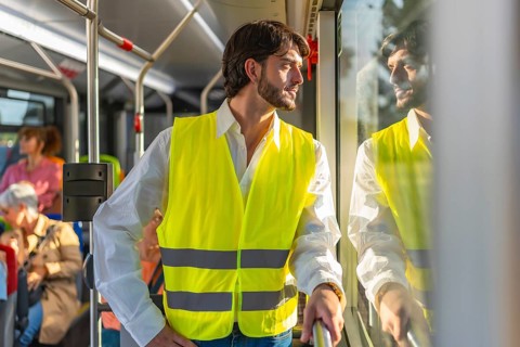 Young man in reflective safety vest standing on a city bus, looking out the window at sunlight and