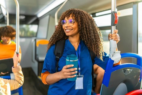 Young black nurse in uniform and backpack stands smiling on a busy city bus, holding a reusable cup