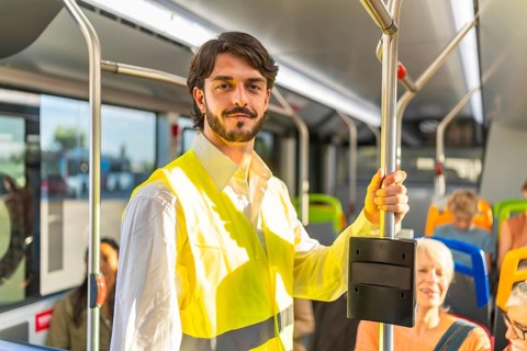 Young man in a high visibility safety vest stands on a modern city bus, holding a pole while