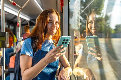 Young woman smiling on a sunny bus commute, holding her smartphone and looking out the window with