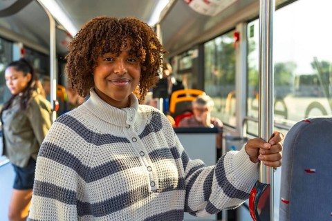 Smiling young woman with curly hair standing on a busy city bus, holding a pole and looking at the