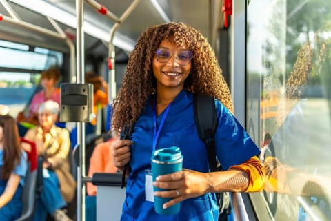 Smiling female nurse or doctor in scrubs and a backpack holding a travel mug, riding public