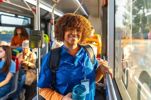 Young nurse in blue scrubs and backpack smiles at camera while standing on a city bus, holding a