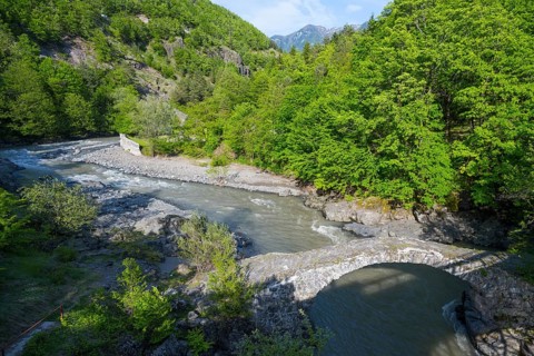 A clear river flows under an old stone bridge through a green, wooded valley with mountains in the