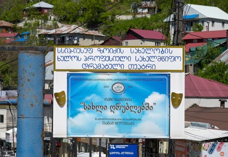 A large advertising sign with a blue poster and text surrounded by colorful buildings and green