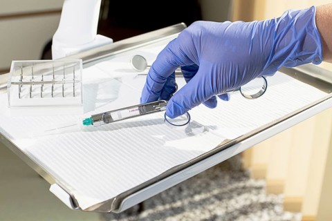 The dentist's hand, in a blue glove, holds a carpal syringe on a tray, ready for a medical