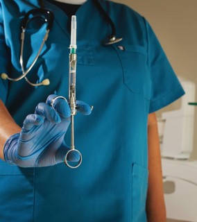 The dentist, in a blue lab coat and gloves, holds a carpal syringe in front of him