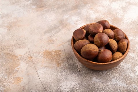 Wooden bowl with raw edible ?hestnuts on brown concrete background, side view, copy space,