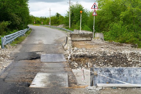 Partially closed road with signs and debris surrounded by green vegetation, bridge over the Techuri