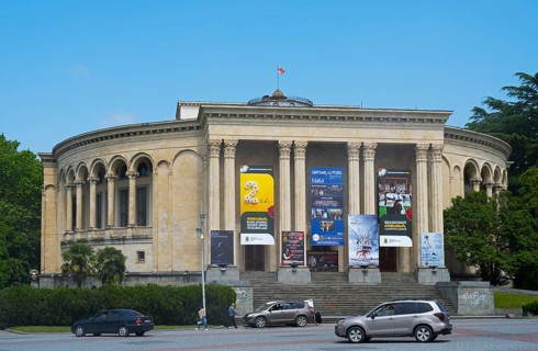 A round classical building with pillars and posters against a blue sky surrounded by green trees